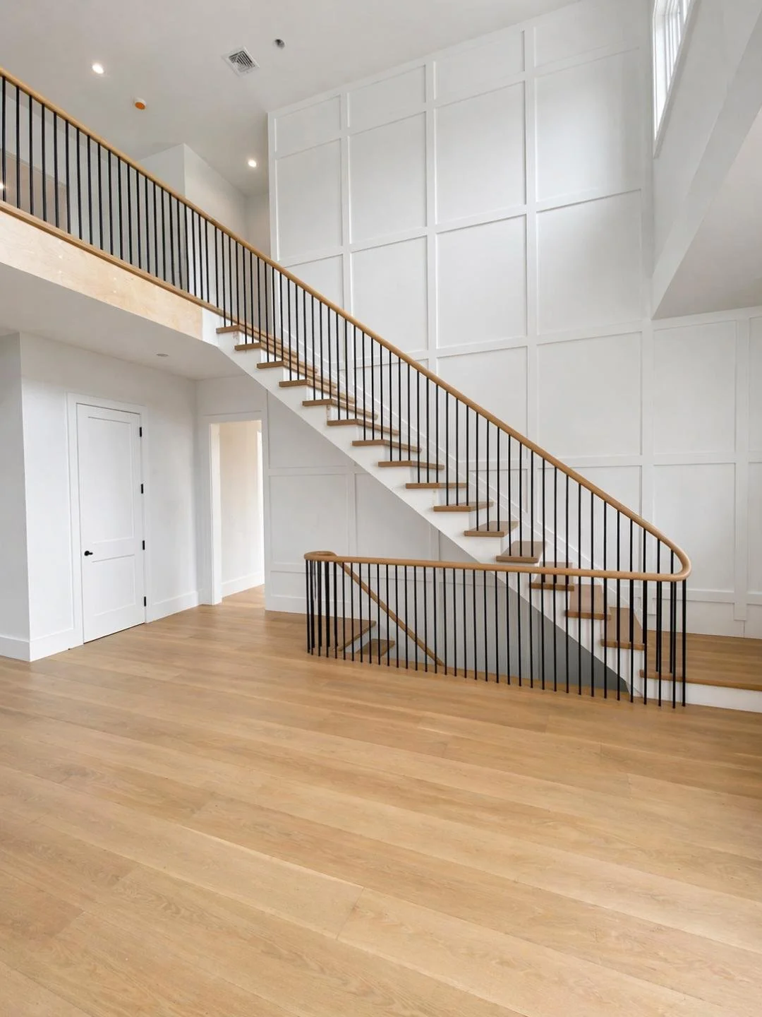 Empty modern indoor staircase with wooden steps, black metal railing, white paneled wall, light wood flooring, and high ceiling with lighting