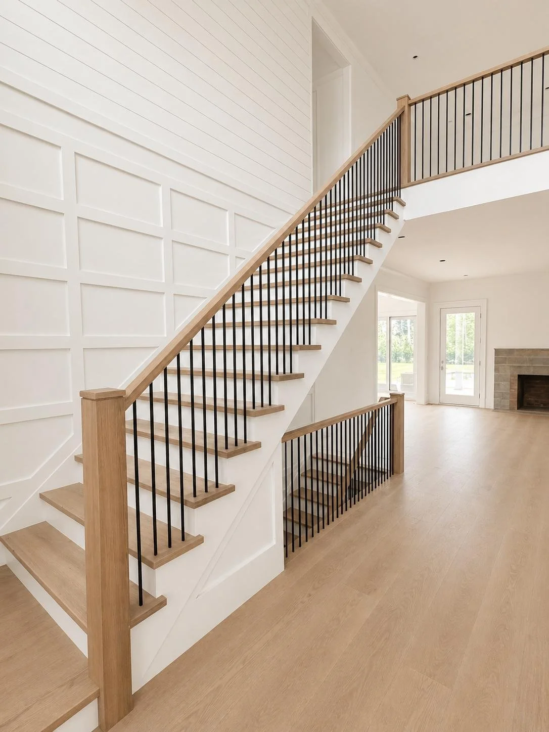 Interior view of a modern staircase with wooden steps, black metal balusters, and white wall paneling in a bright living space with large windows and a fireplace.