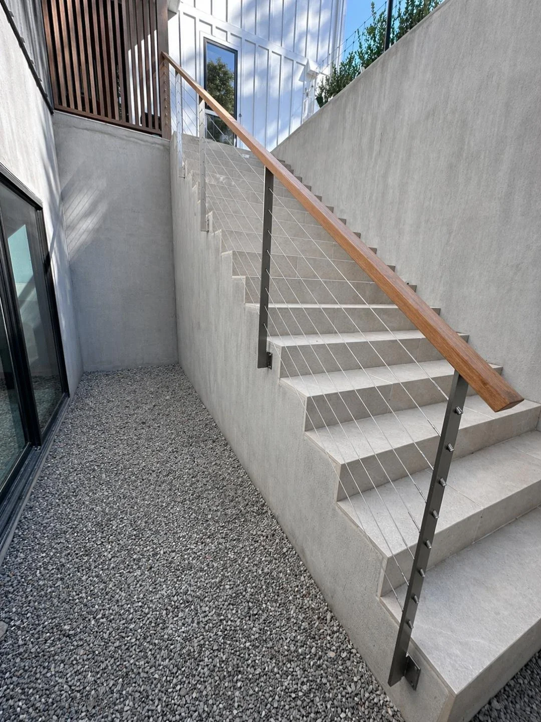 Outdoor stairs with wooden handrail and wire cable railing next to a gravel path, leading up to a modern house.