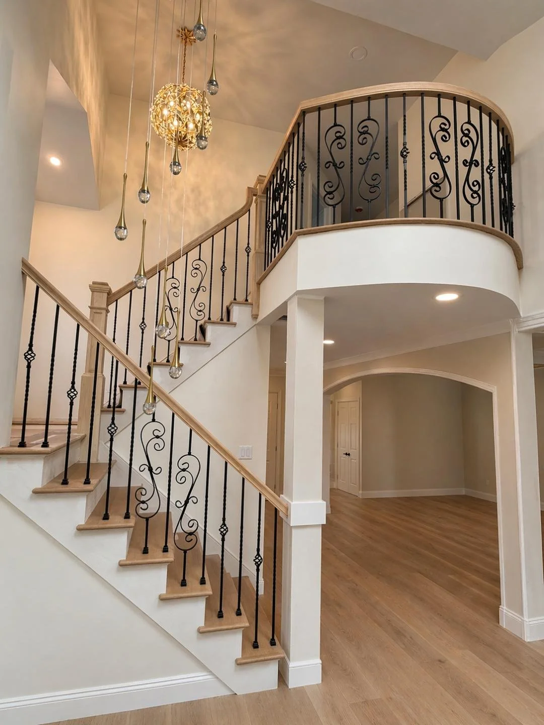 Interior of a house with a staircase and second-floor balcony, featuring decorative wrought iron railings, wooden steps, hardwood flooring, and modern hanging light fixtures.