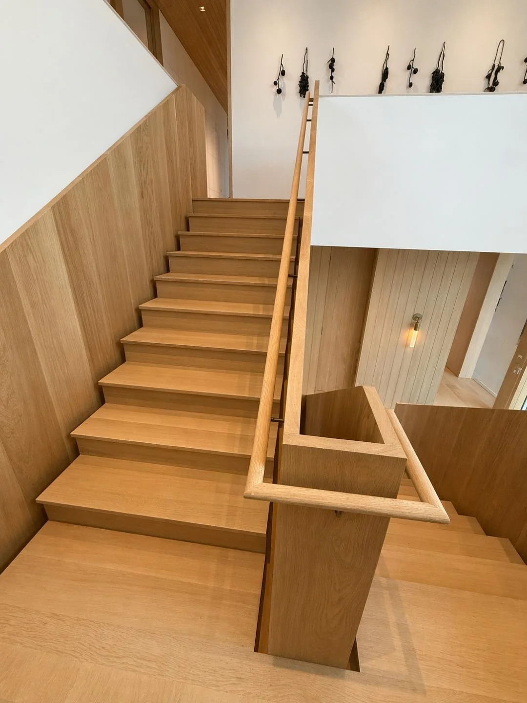 Wooden staircase with minimalist design in modern interior, featuring wooden panels on the wall and a white wall with hanging black abstract art or decorative elements.