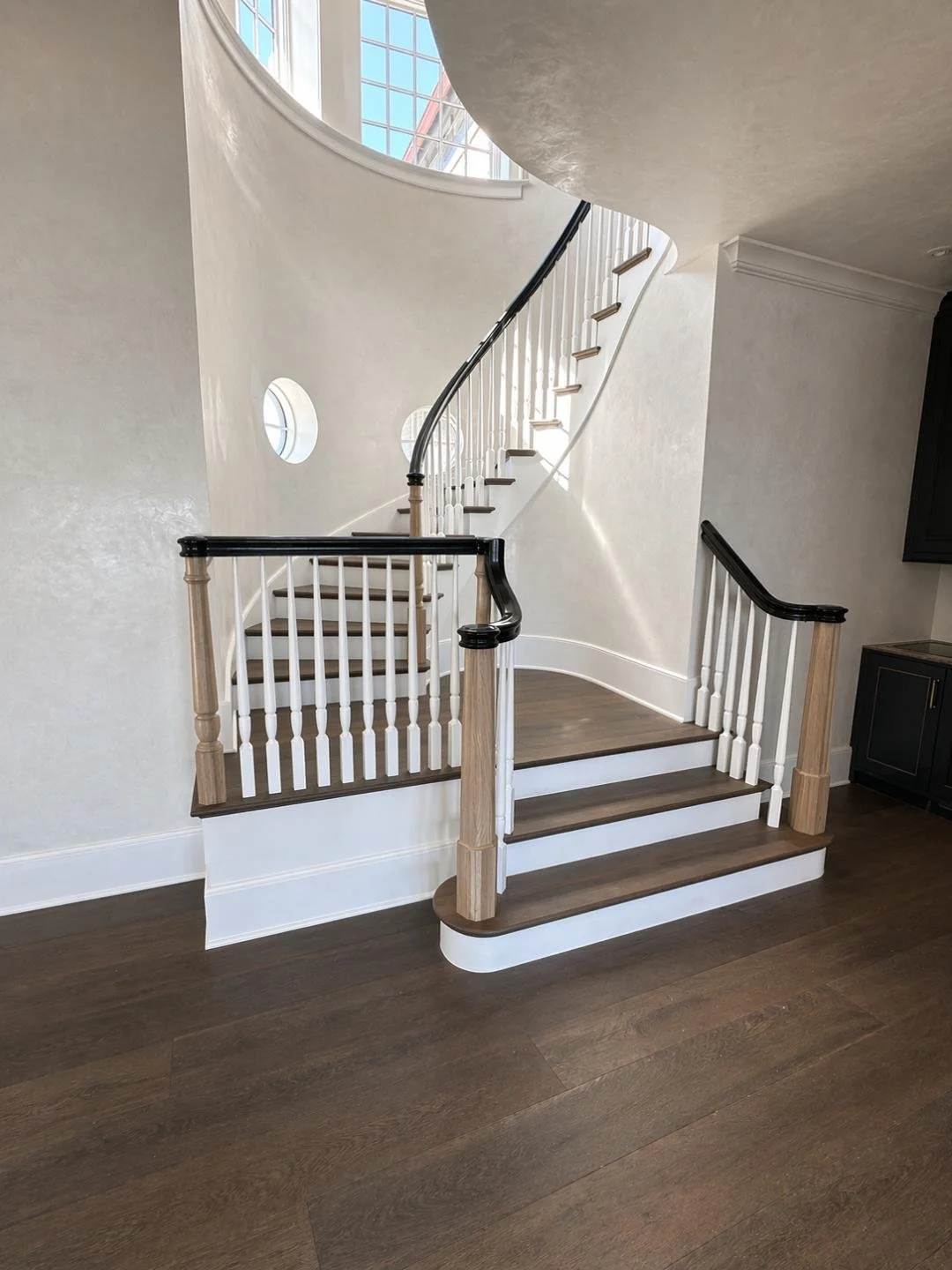 Interior view of a curved staircase with wooden steps, white spindles, black handrail, and a large window letting in natural light.