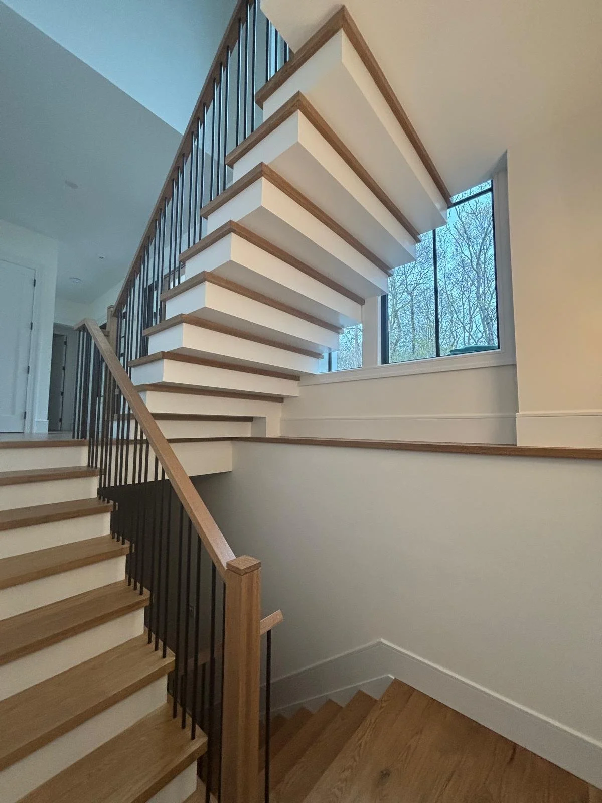 Inside view of a staircase with wooden steps, black metal railing, and a large window showing trees outside.