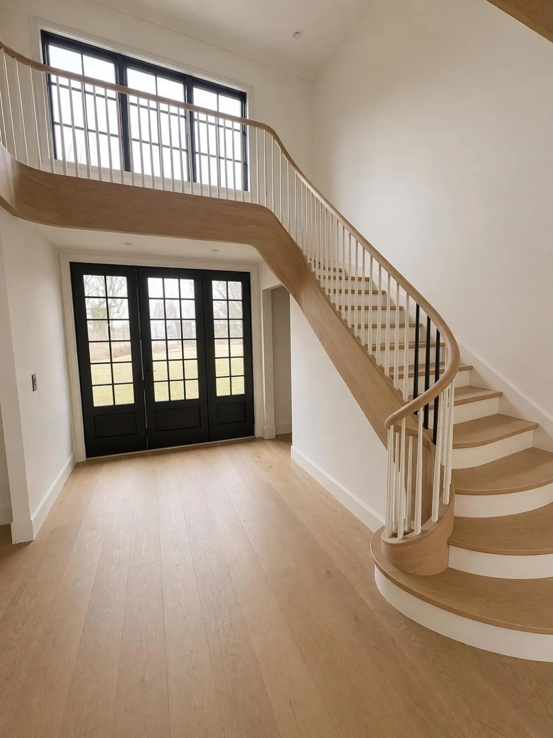 Interior view of a modern house foyer with a curved wooden staircase, black double doors with glass panels, and large windows letting in natural light. The floor is light wood, and the walls are white.