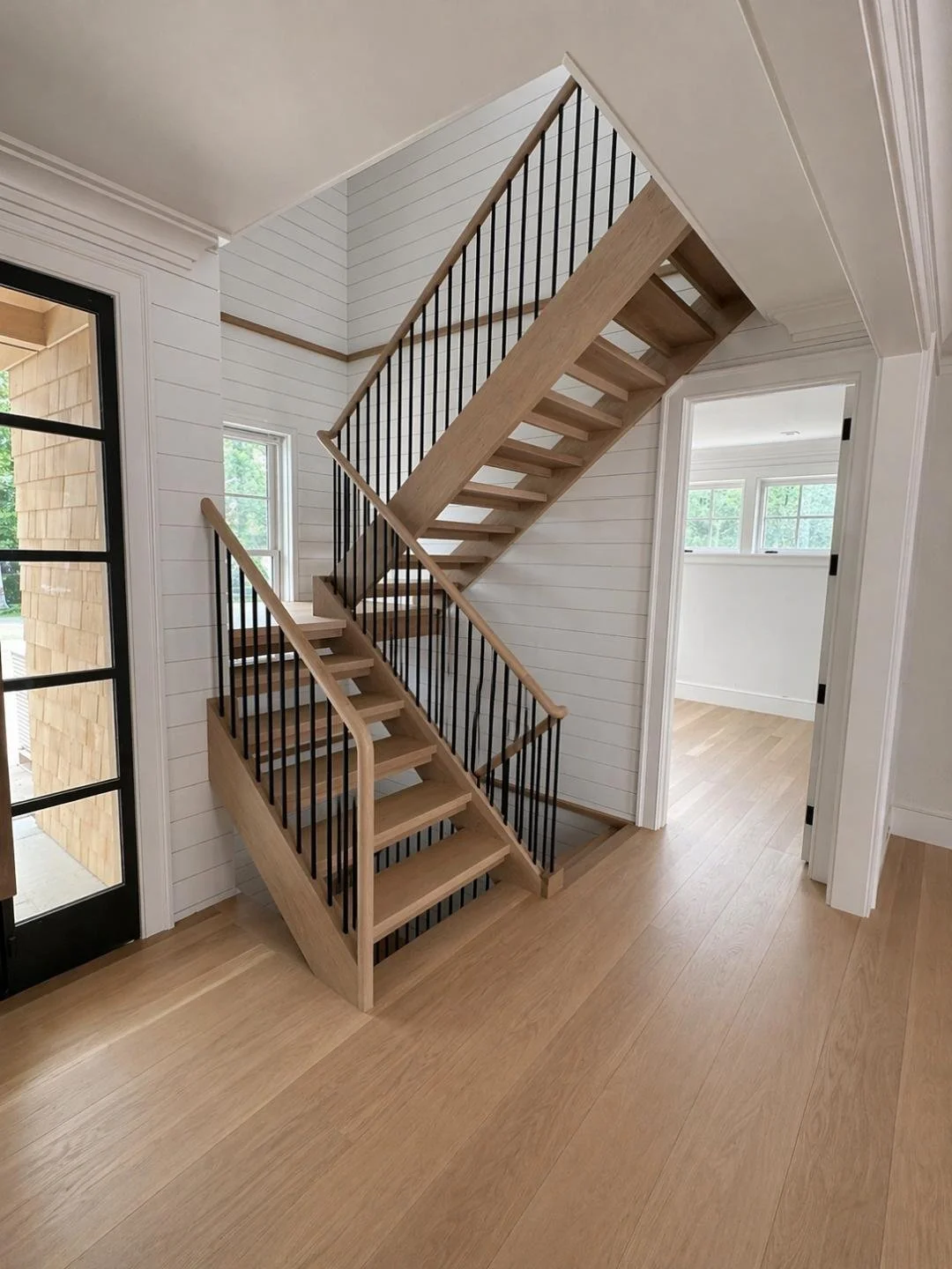 Interior view of a house with a wooden staircase with black metal spindles, white shiplap walls, and light wooden flooring, leading to upper and lower levels.