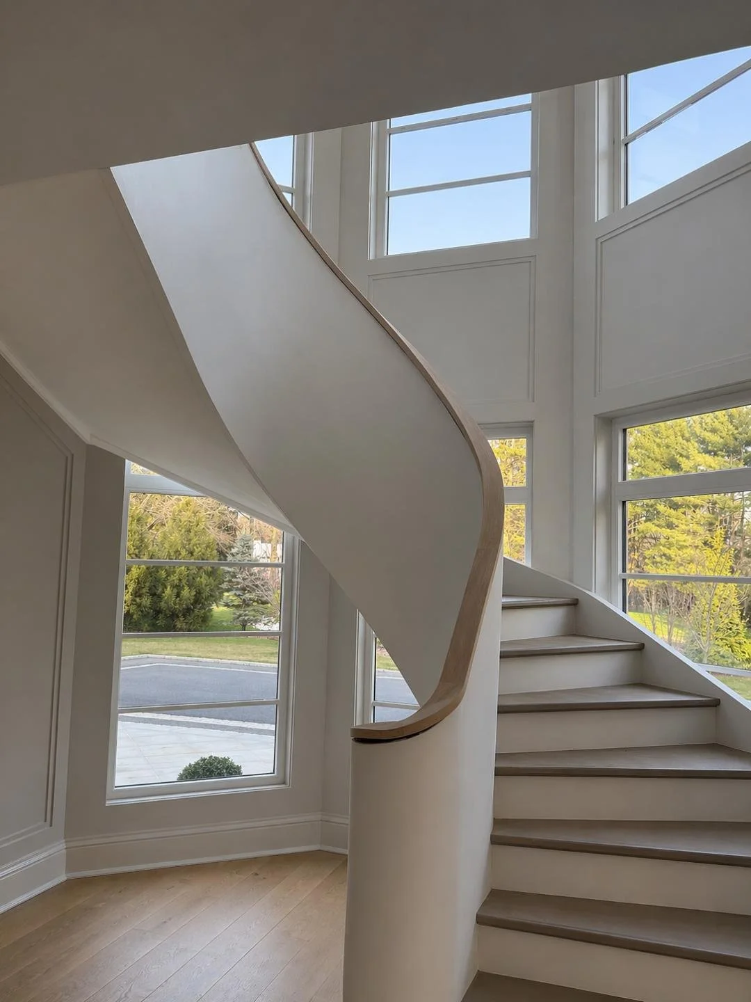 Interior view of a modern staircase with a curved wooden handrail, white walls, and large windows letting in natural light, overlooking a garden.