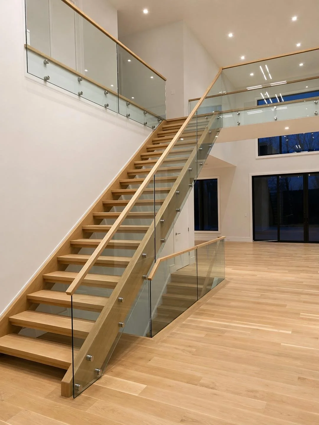 Empty modern interior featuring wooden staircase with glass railing, large windows, and hardwood flooring.