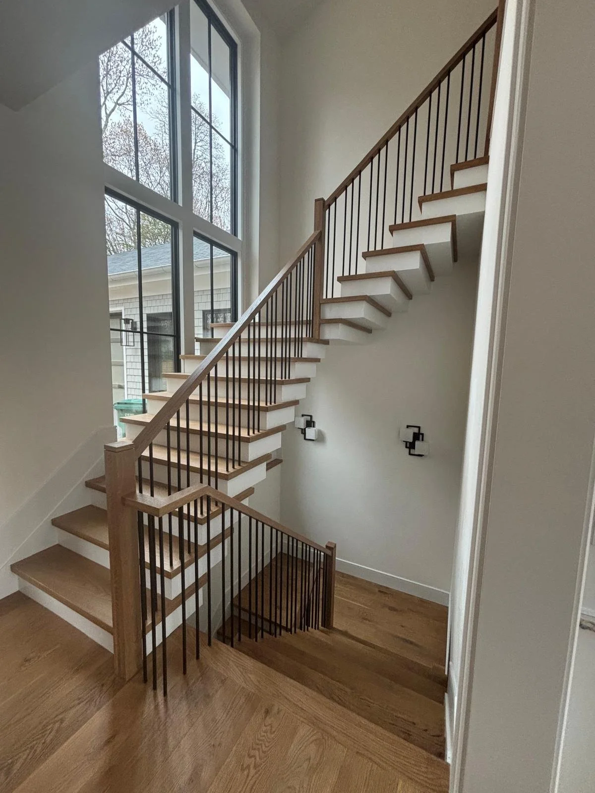 Wooden staircase with black metal railings inside a house, leading to an upper floor, next to large windows letting in natural light.