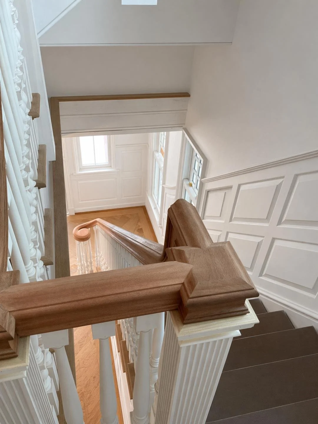 View from the top of a staircase looking down, featuring wooden handrails, white columns, and walls with decorative paneling, in a well-lit home interior.