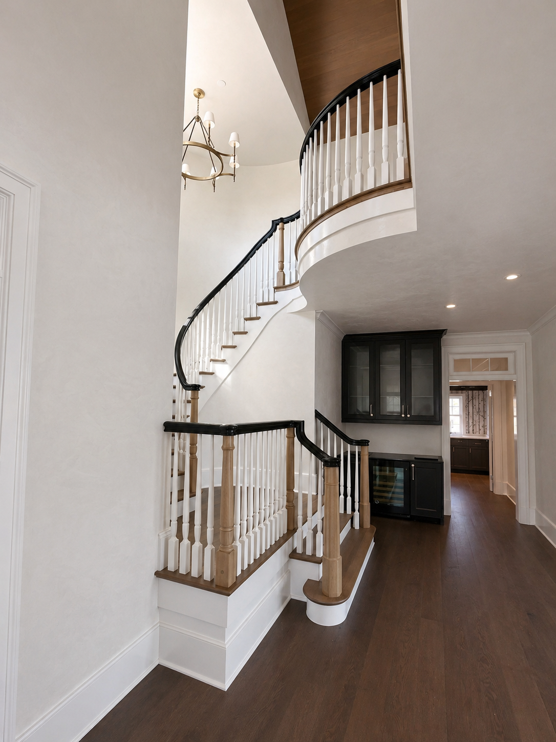 Interior view of a home staircase with a curved dark wooden handrail, white spindles, and wooden steps, leading to an upper level. Dark wood cabinets and a doorway are visible in the background.