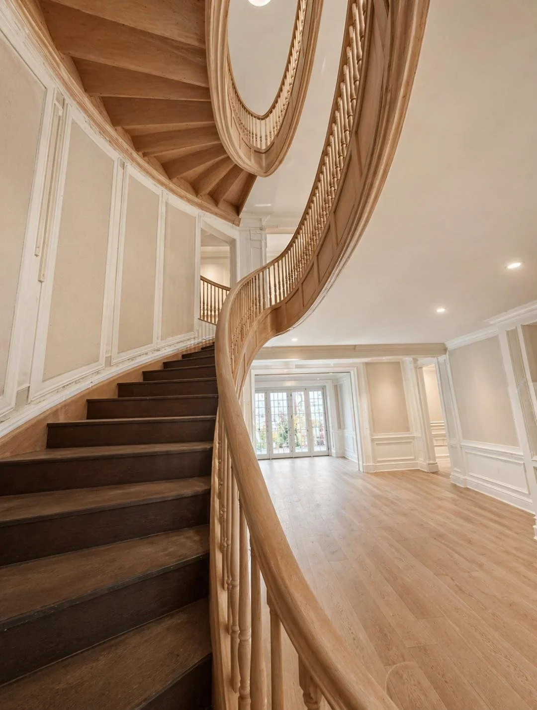 Interior view of a grand staircase with wooden railing and dark stairs, curved upwards in a spacious room with hardwood floors and white paneled walls, large windows at the far end.