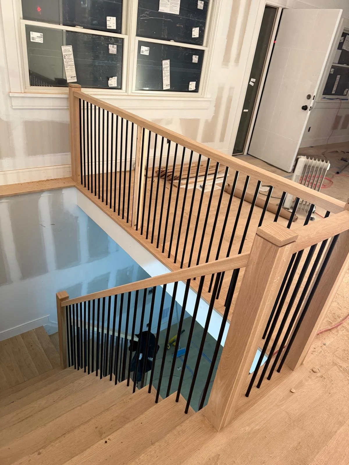 Wooden staircase with black metal balusters surrounding a cut-out opening for a lowered section, leading to a lower floor under construction in a house.