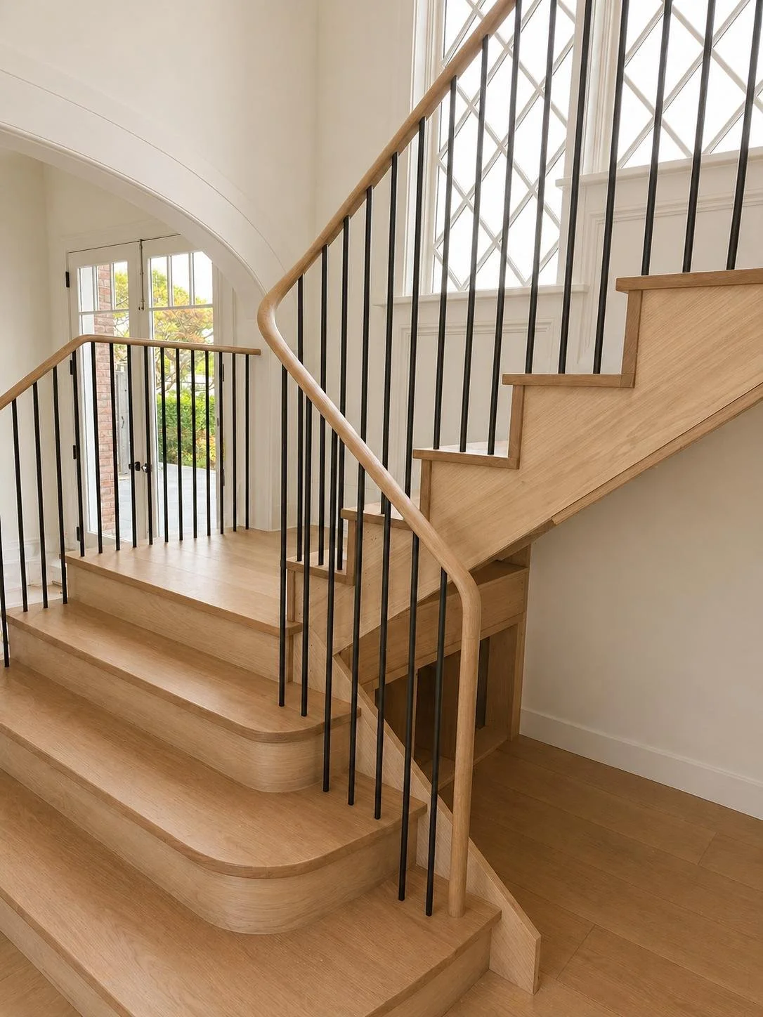 Wooden staircase with black metal balusters and a curved handrail, near large windows with diamond-patterned grid, in a bright, modern interior.