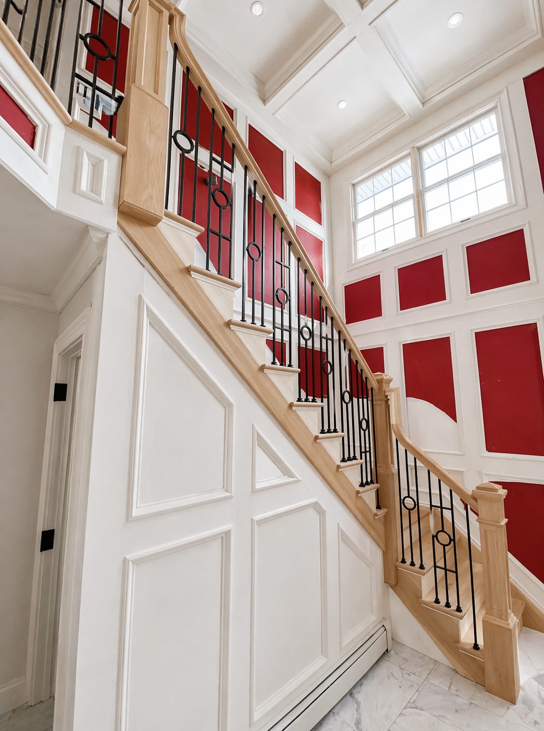 Interior view of a staircase with wooden handrail and black metal balusters, white wainscoting, and red paneled walls near large windows.
