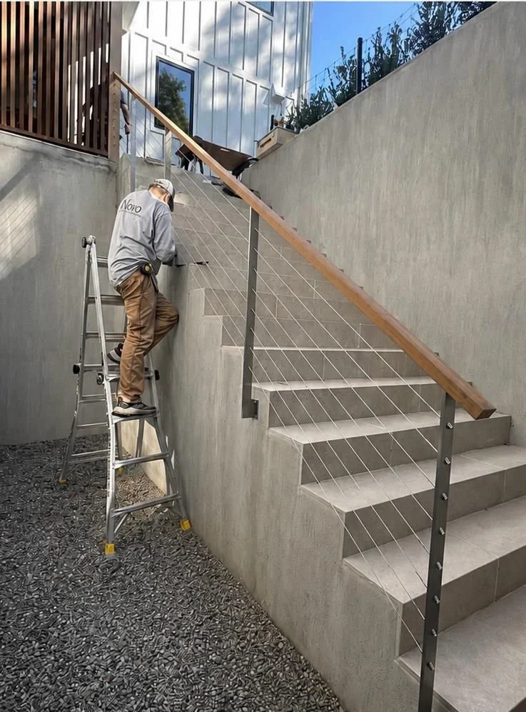A worker on a ladder installing a cable on a staircase railing outdoors, with a modern building in the background.