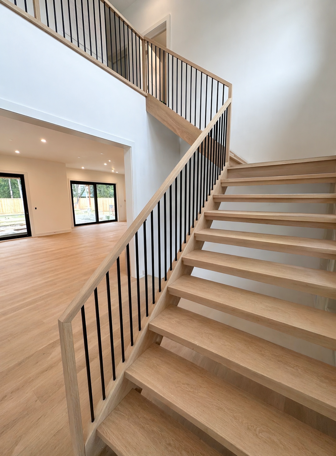 Interior of a modern house under construction or renovation, featuring a wooden staircase with black metal balusters and light wood flooring, leading to an upper level with a white wall and an open living area with sliding glass doors.