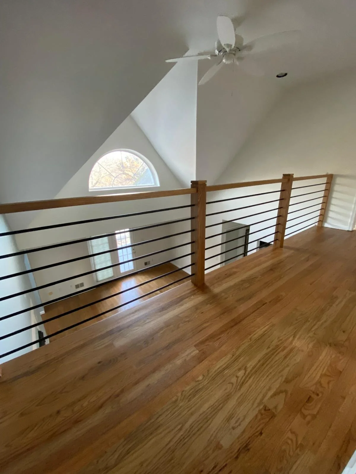 An interior view of a second-floor balcony with a wooden floor and black metal railing, overlooking a room with a large arched window and high vaulted ceiling with a ceiling fan.