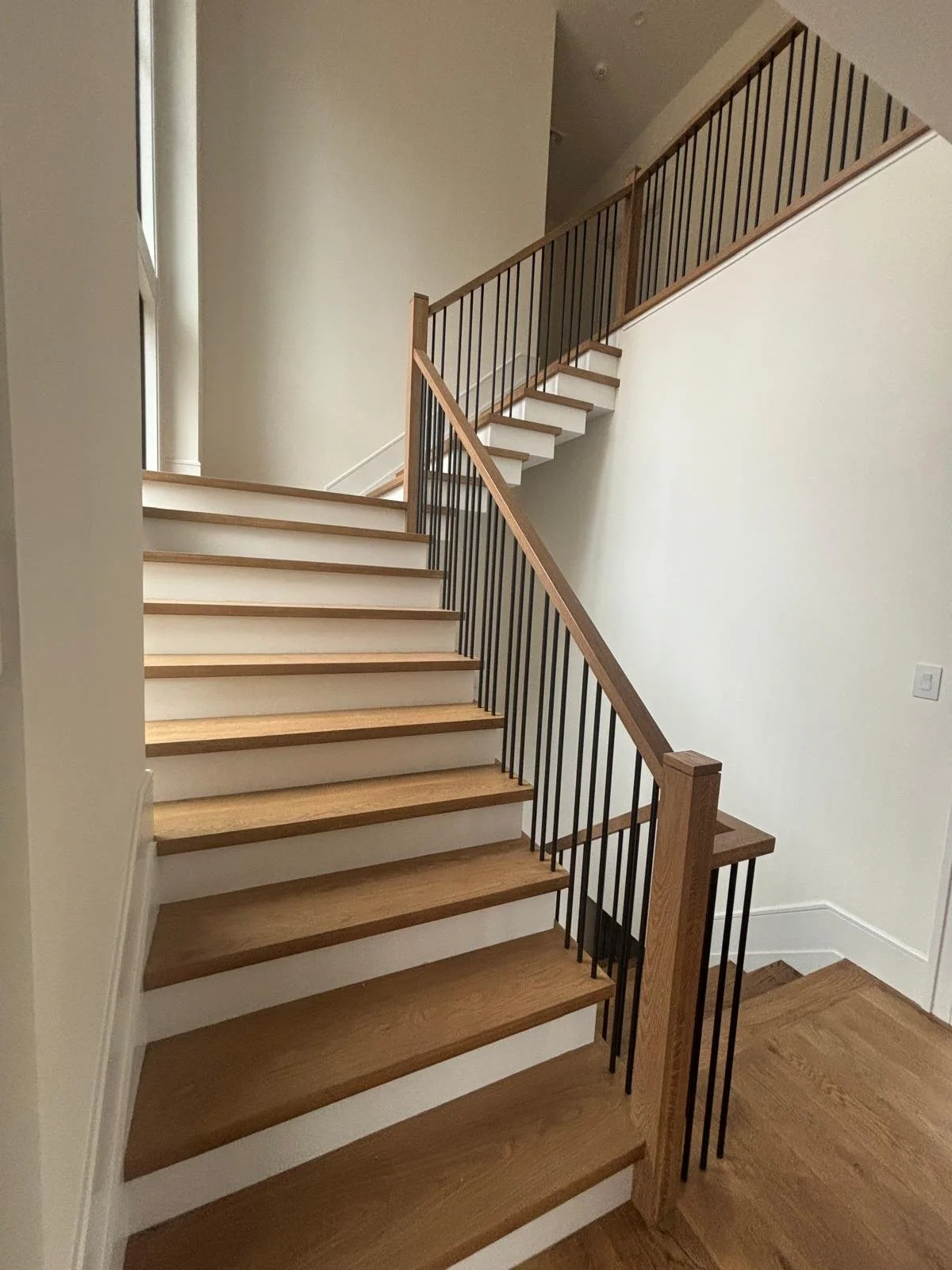 Wooden staircase with black metal railing in a modern home, leading up to an upper floor with light-colored walls and large windows.