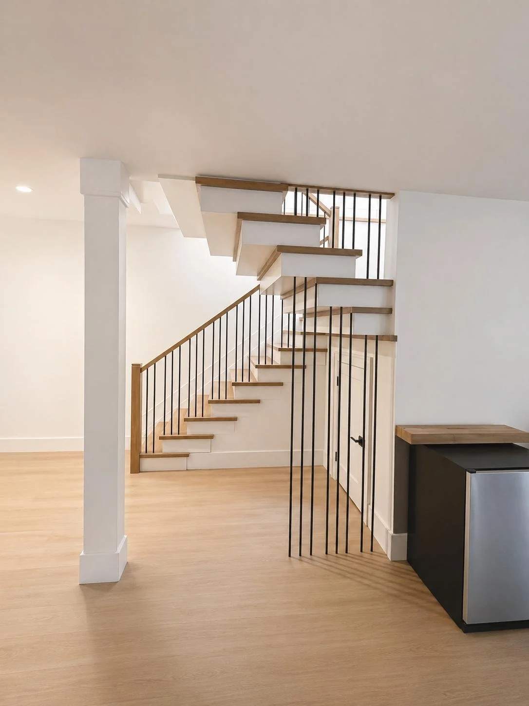 Modern staircase with wooden steps and black vertical railings in a minimalist interior with light wood flooring and white walls.