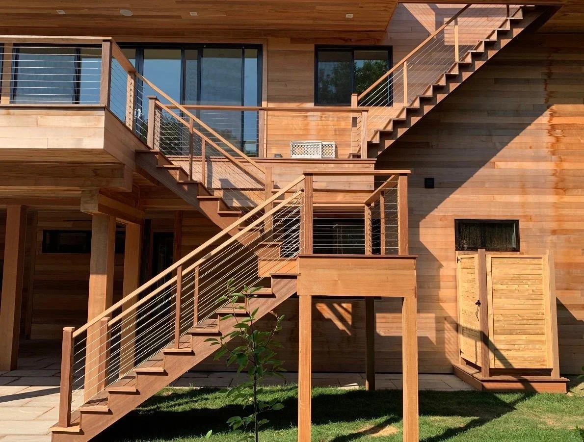 Wooden multi-level outdoor staircase and deck of a modern house with large glass windows, surrounded by grass and sunlight.