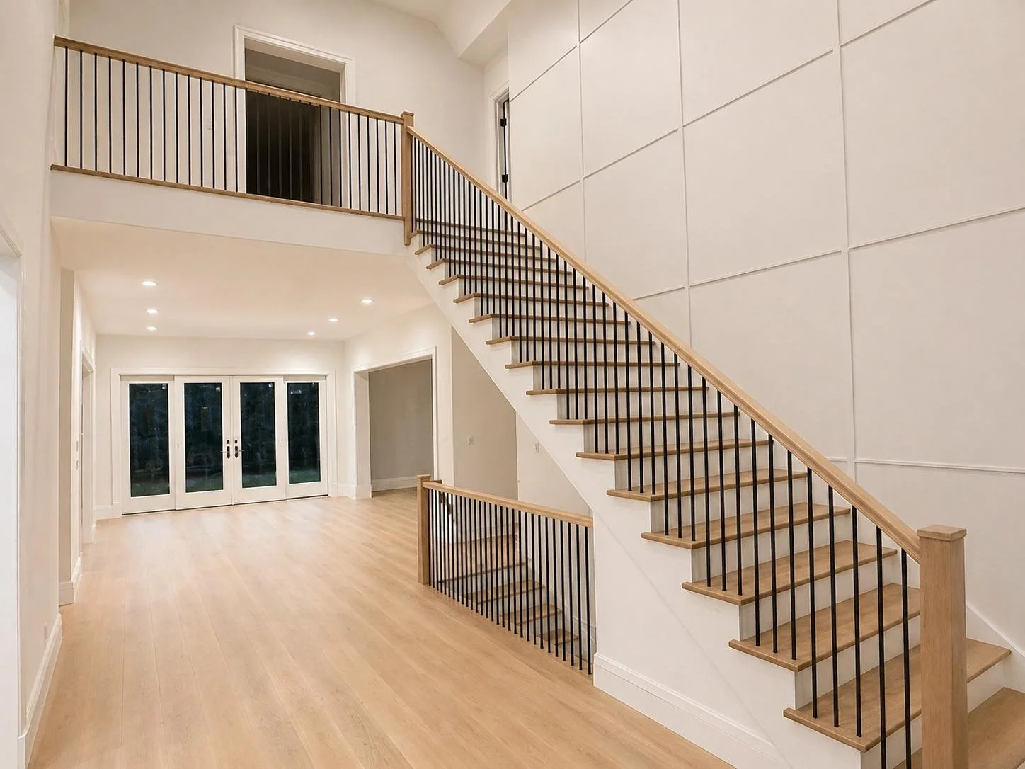 Empty living area with wooden flooring and a staircase with wooden steps and black metal railings leading to an upper level. Double glass doors at the far end.