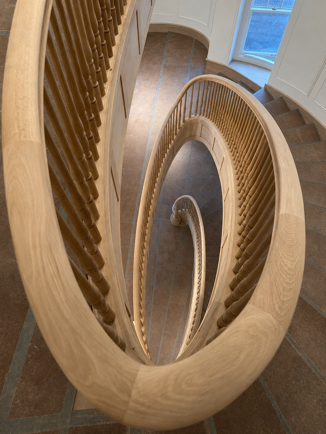 Spiral staircase with wooden handrail and balusters, viewed from above, in a home interior with tiled flooring and a large window.