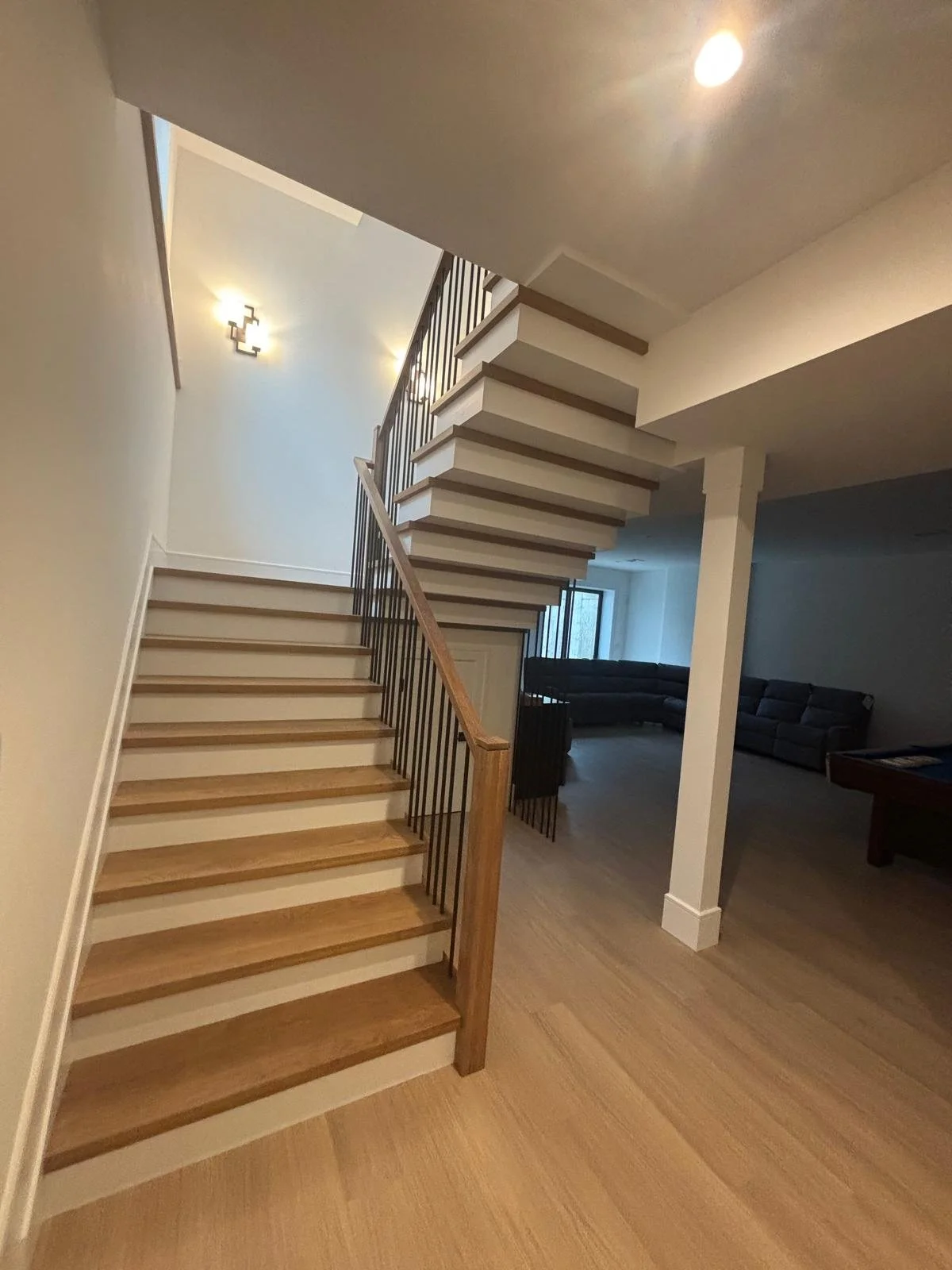 Interior view of a modern staircase with wooden steps and metal railings, leading to an upper floor, with a living room visible in the background.