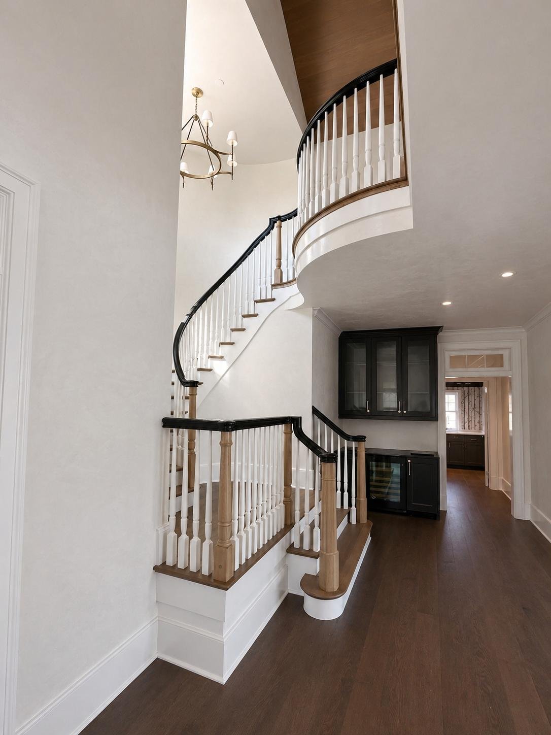 Interior view of a staircase with black and natural wood railing, leading to a second floor, with dark wood cabinets and hallway in the background, white walls, and hardwood flooring.
