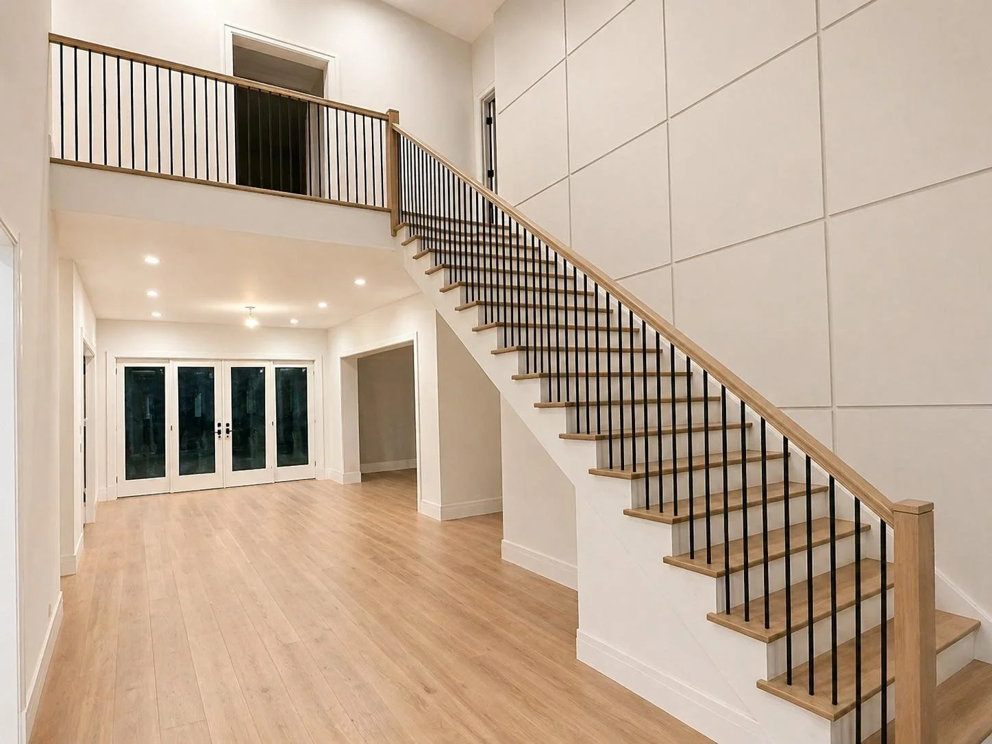 Empty living space with wooden flooring, a staircase with wooden steps and black metal balusters, and a glass door at the back leading outside.