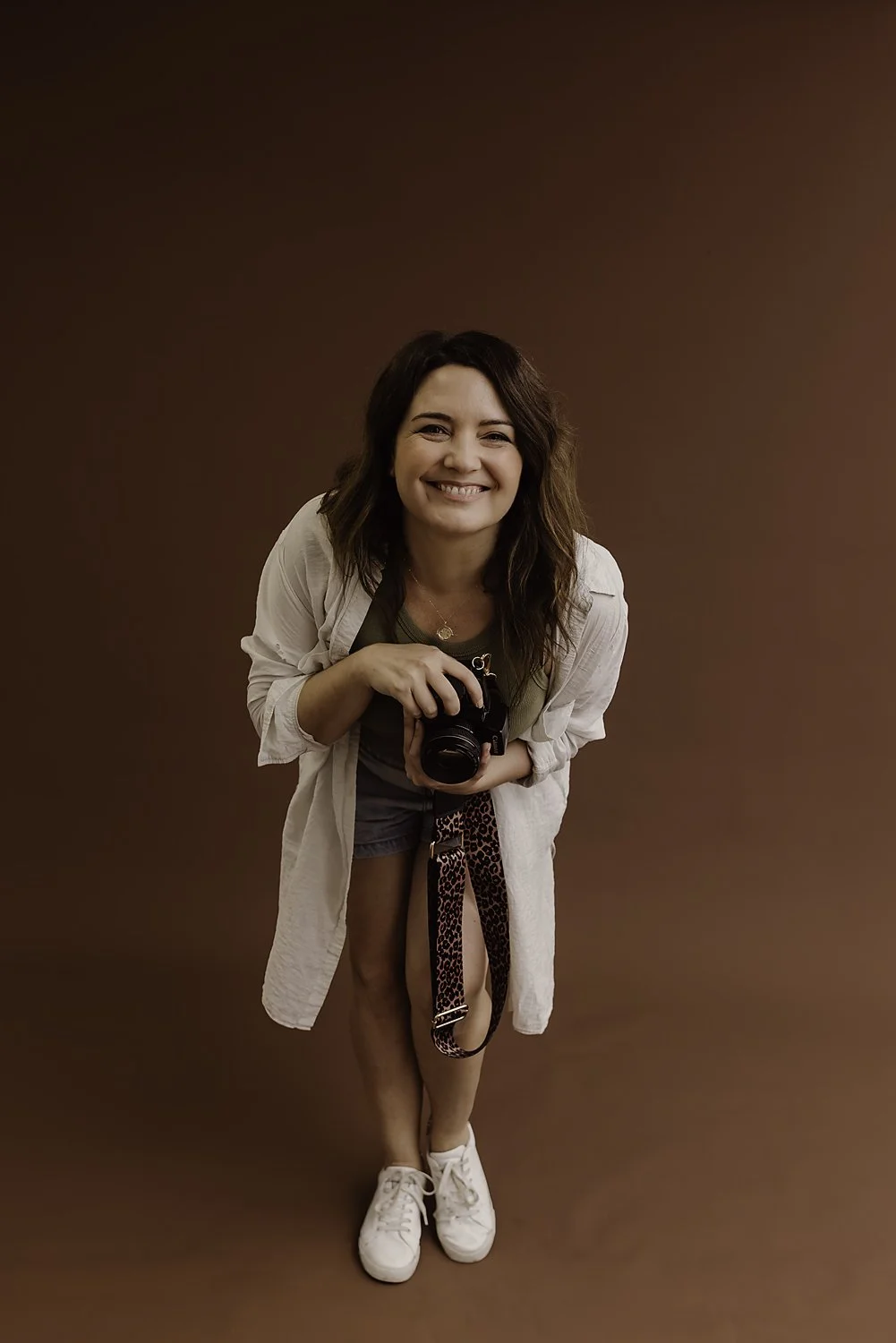 Woman smiling, holding a camera, wearing a white shirt and sneakers, standing on a brown background.