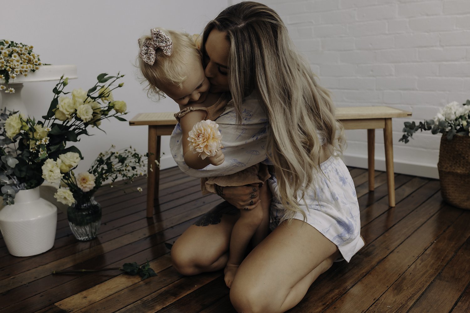 Woman kneeling and holding a toddler who is holding a flower, surrounded by vases of flowers on a wooden floor.