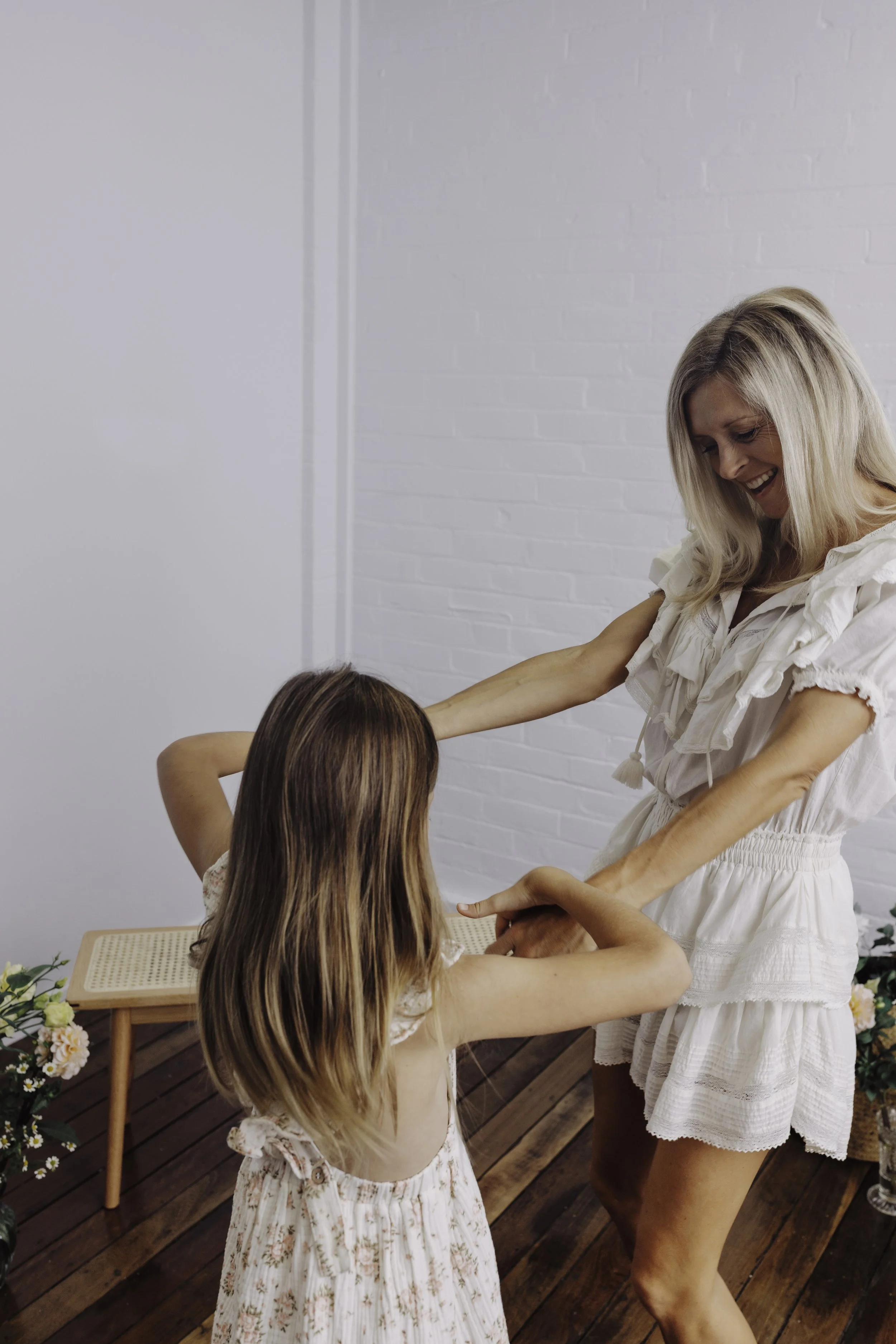 Woman in white dress dancing with young girl in floral dress on wooden floor, white brick wall in background.