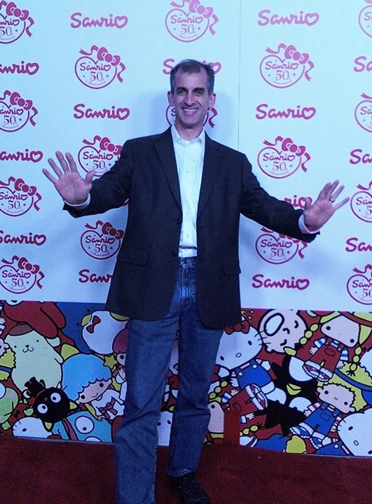 Man in a blazer and jeans smiling with arms outstretched, standing in front of a Sanrio-themed backdrop with characters and logos.