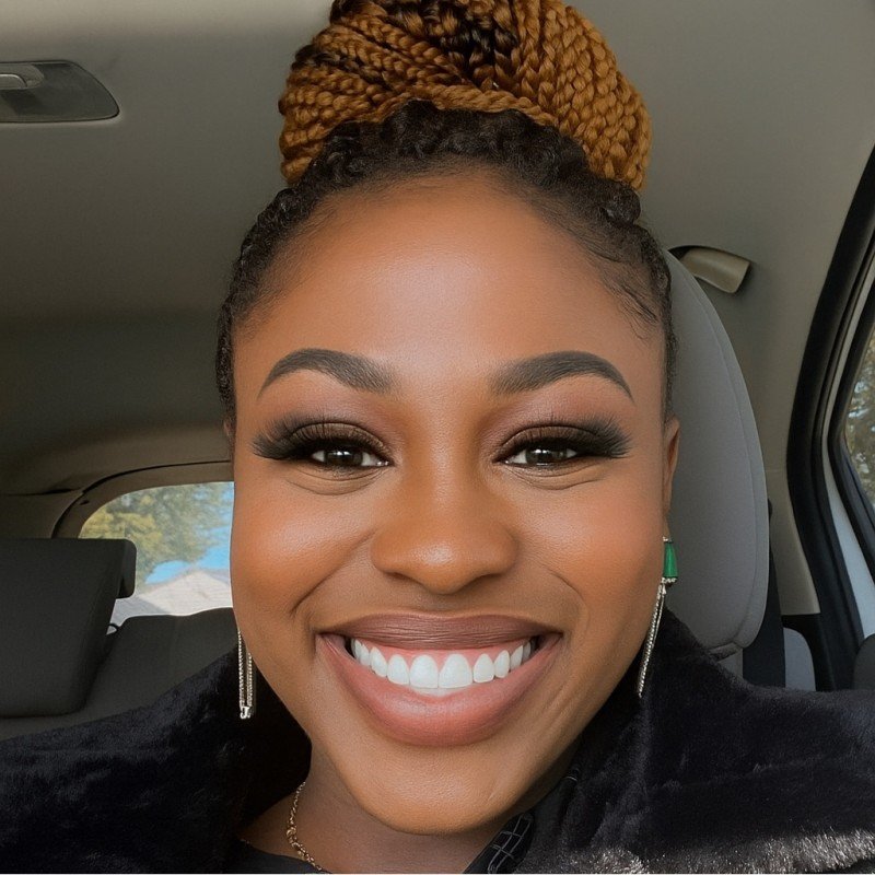A young woman with long, straight, reddish-brown hair taking a selfie against a plain white wall. She is smiling and wearing a black turtleneck sweater.
