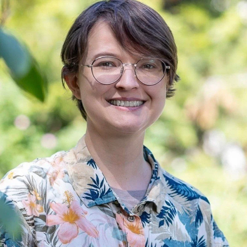 A woman with short brown hair and glasses, smiling outdoors in a garden, wearing a floral shirt.
