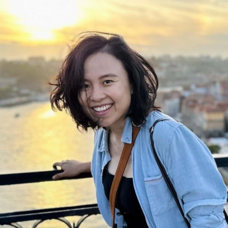 A woman with shoulder-length dark hair smiling at the camera, wearing a light blue denim jacket outdoors during sunset with a cityscape and water in the background.