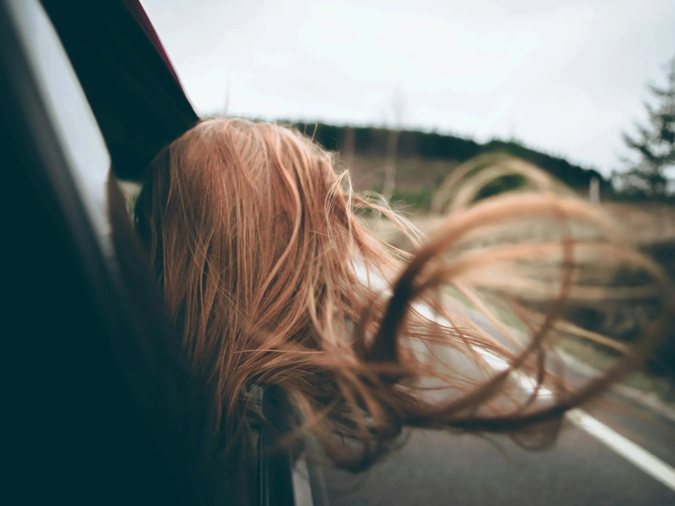 A person with long, wavy red hair leaning out of a vehicle window on a road with a landscape of hills and trees in the background.