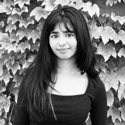 A young woman with long dark hair standing in front of a background of leafy plants, smiling at the camera.
