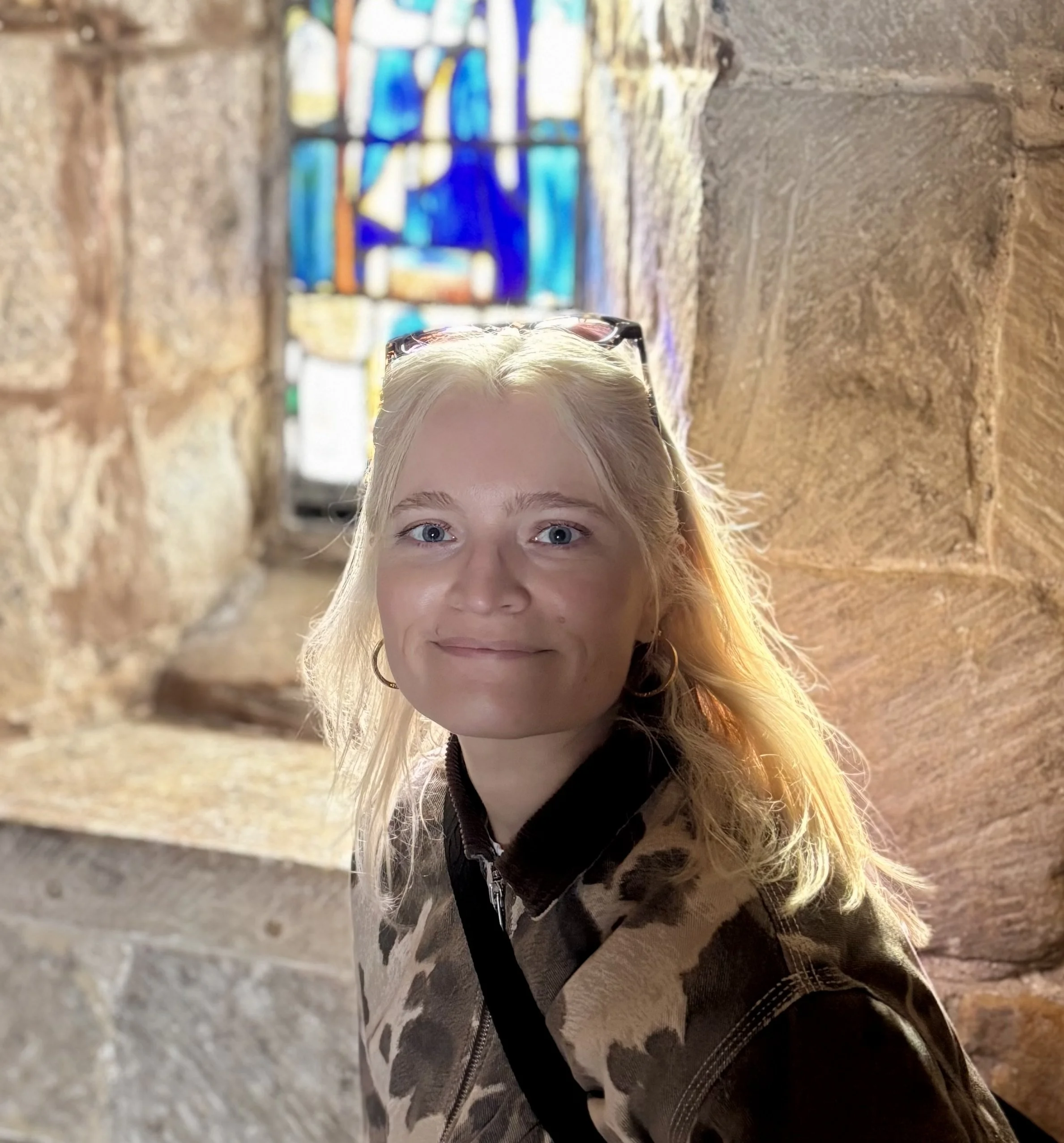 A young woman with blonde hair and blue eyes poses inside a stone building with stained glass window behind her.