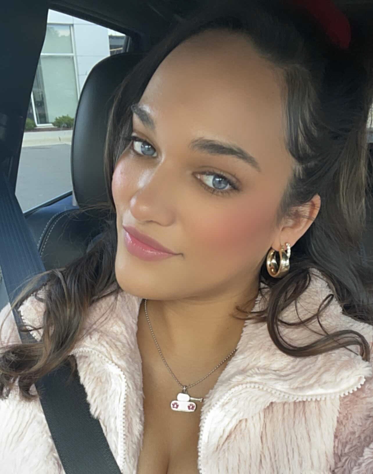 A woman with long brown hair and blue eyes, wearing gold hoop earrings and a tank-themed necklace, sitting in a car with a seatbelt on, smiling.