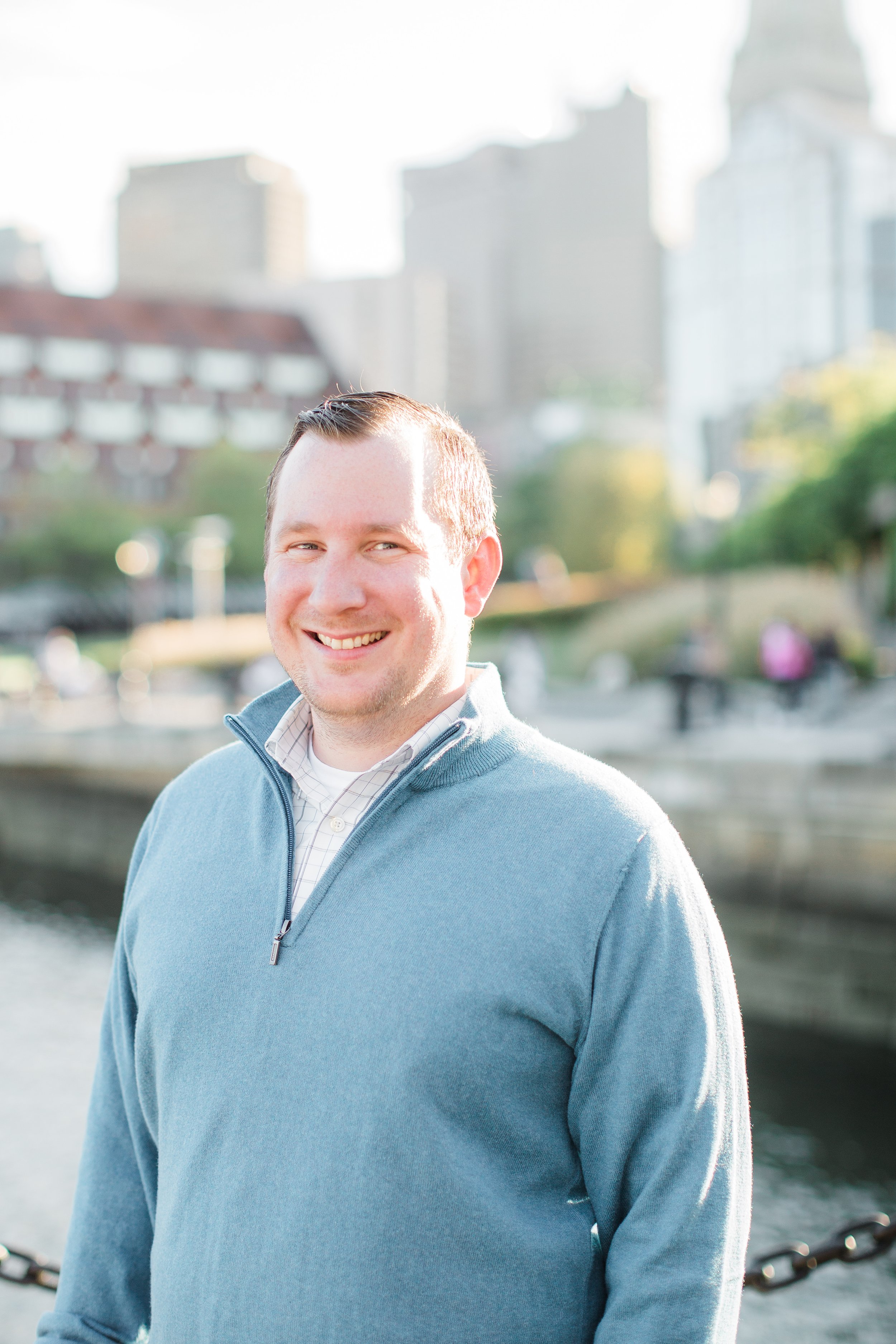 A smiling man with short hair wearing a light blue quarter-zip sweater and a collared shirt stands outdoors near a waterway with city buildings in the background.