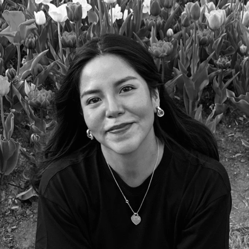 A woman with long dark hair and earrings, smiling, sitting in front of a flower garden.