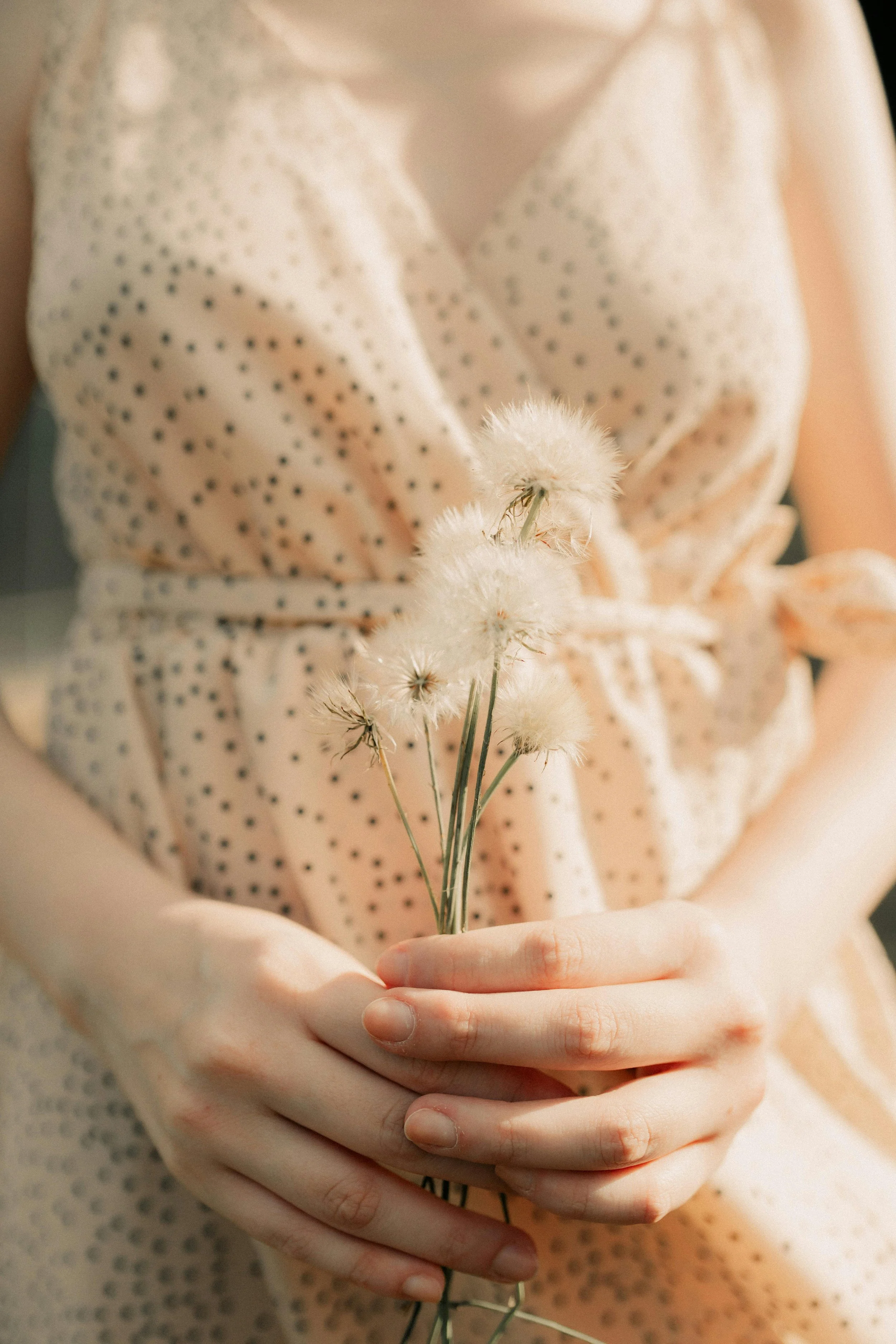 A person wearing a light-colored, polka-dot dress holding a small bunch of white fluffy flowers.