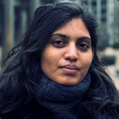 Close-up of a young woman with dark, wavy hair wearing a dark scarf against an urban background.