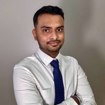 A man in a white shirt and blue tie, with arms crossed, posing against a neutral background.