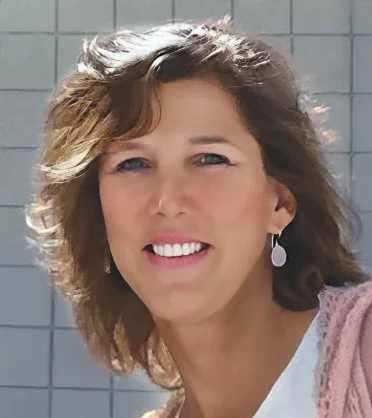 A smiling woman with shoulder-length brown hair, wearing a white top and a pink cardigan, standing outdoors with a grid tile background.