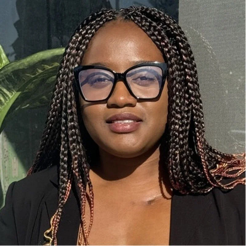 A woman with braided hair wearing large black glasses and a black jacket, standing outdoors near a large green plant.