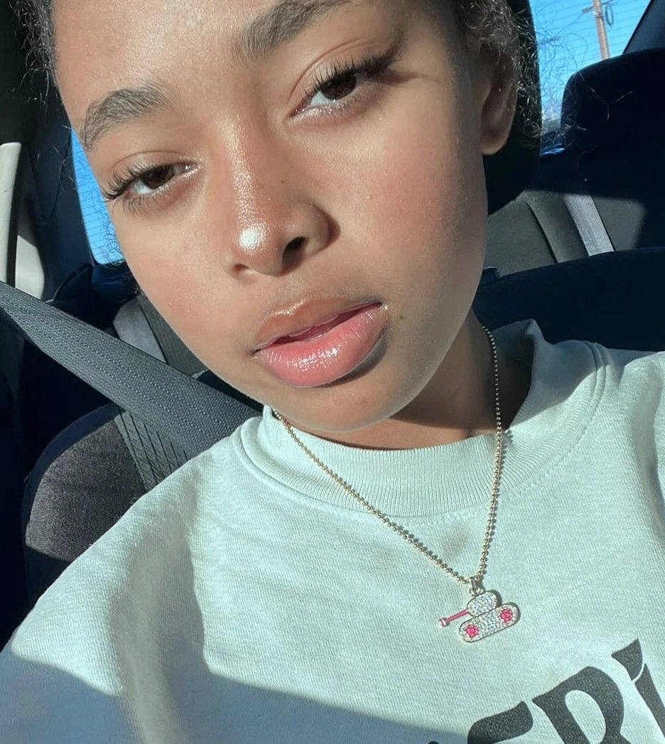 Close-up of a young woman with light brown hair and makeup, wearing a white shirt and a necklace with a tank charm, sitting in a car with sunlight on her face.