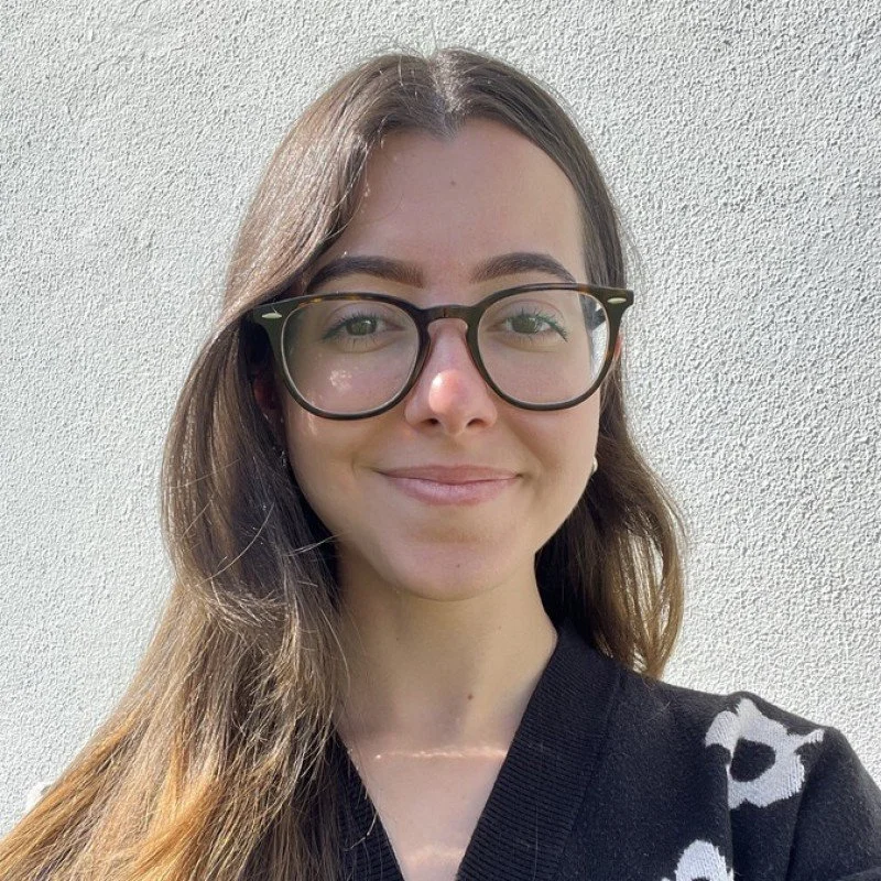 A young woman with long brown hair and glasses, smiling in front of a textured white wall.