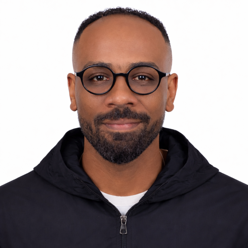 A man with salt-and-pepper beard wearing round glasses and a black zip-up jacket against a white background.